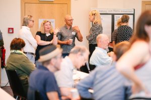 Eine Gruppe von Personen die sich unterhält. Im Vordergrund sind weitere sitzende Personen abgebildet.