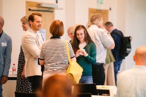 Eine Gruppe von Personen die sich unterhält. Im Vordergrund sind ein Mann und zwei Frauen, die sich unterhalten und lächeln.