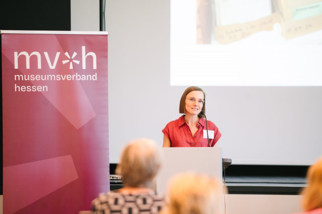 Dr. Saskia Johann vom Museumsverband Hessen steht an einem Rednerpult mit Mikrofon. Im Hintergrund ist ein dunkelroter Banner des Museumsverbandes mit Logo und eine Leinwand zu sehen.