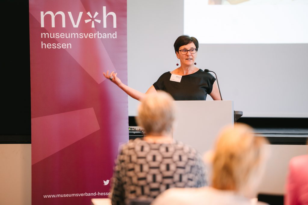 Vera Kudlinski vom Museumsverband Hessen steht an einem Rednerpult mit Mikrofon und spricht Richtung Publikum. Im Hintergrund ist ein dunkelroter Banner des Museumsverbandes mit Logo und eine Leinwand zu sehen.