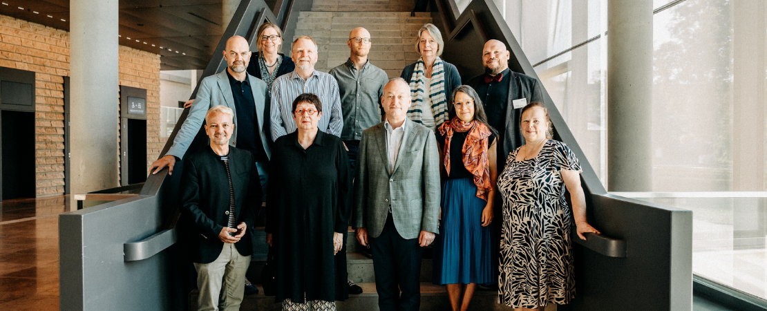 Gruppenfoto von elf Personen auf einer Treppe in einem modernen Gebäude. Die Personen stehen in zwei Reihen, blicken in die Kamera und lächeln. Im Hintergrund sind Glasfassaden, Betonwände und Holzverkleidung zu sehen.