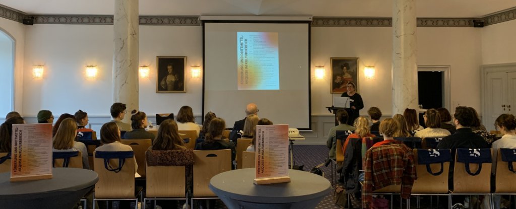 Vortragssituation in einem historischen Saal: Eine Referentin spricht vor einem sitzenden Publikum, im Hintergrund eine Leinwand mit Präsentation, umgeben von Säulen und Wandporträts