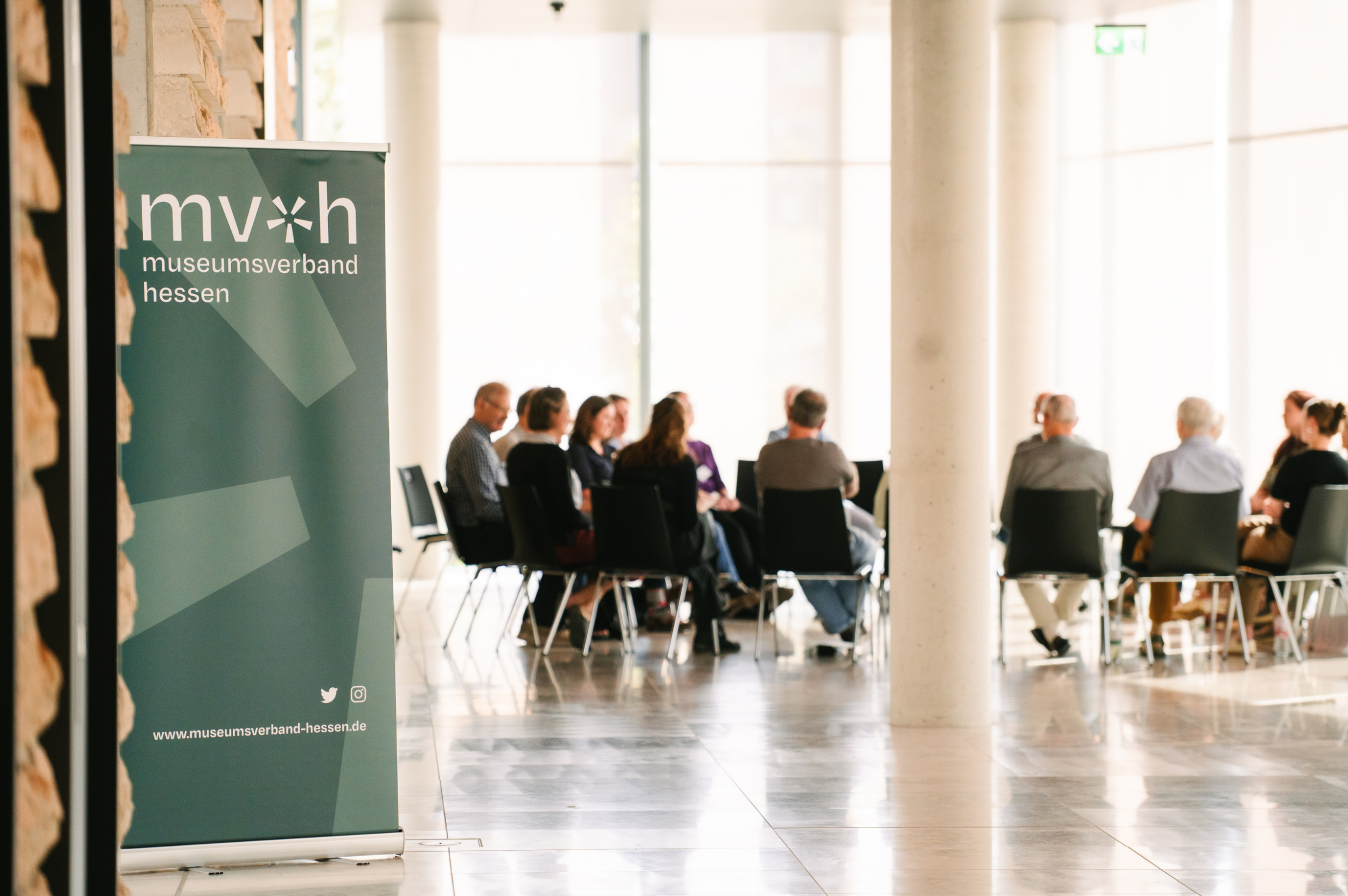 Eine Gruppe von Menschen sitzt in einem hellen Raum in einem Stuhlkreis und diskutiert; links im Bild steht ein Banner des Museumsverbands Hessen.