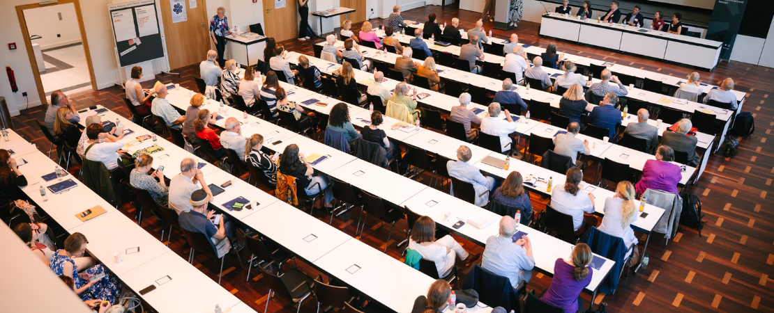 Blick von oben in Veranstaltungsaal, die Reihen sind gut besetzt mit Menschen, vorne eine Leinwand