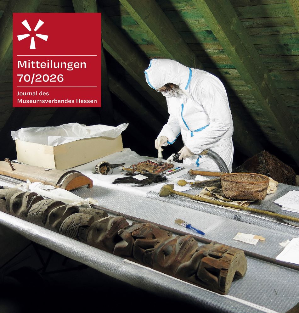 Auf einem Tisch liegen diverse ethnologische Objekte sowie Verpackungsmaterial. Eine Person in einem weißen Schutzanzug säubert mit Staubsauger und Pinsel ein Objekt. Links oben ist ein rotes Feld mit dem Logo des Museumsverbandes Hessen und der Aufschrift: „Mitteilungen 69/2025 – Journal des Museumsverbandes Hessen“.