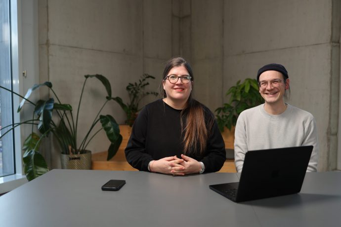 Anne-Marie Bernhard, links und Fabian Fess, rechts, sitzen an einem Tisch in einem modernen Büro mit Betonwänden und mehreren grünen Zimmerpflanzen im Hintergrund. Anne-Marie Bernhard trägt einen schwarzen Pullover und eine Brille, Fabian Fess einen hellen Pullover, eine dunkle Mütze und sitzt vor einem Laptop.