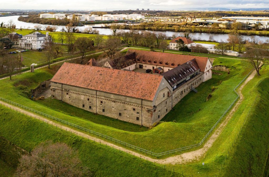 Vierseitige Festung umringt von einem begrünten Festungsgraben. Im Hintergrund befindet sich der Main.
