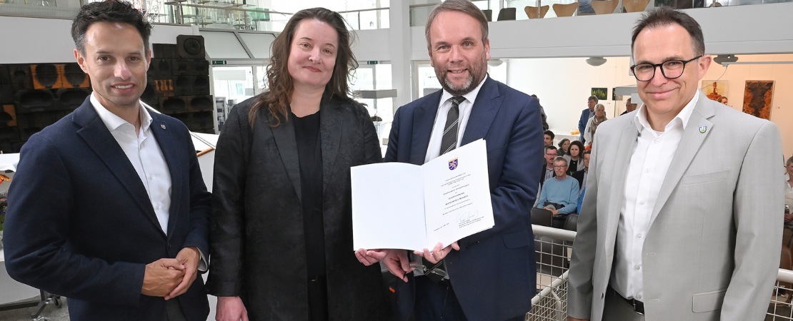 Vier Personen stehen in einem hellen Museumsfoyer bei einer offiziellen Übergabe. Ein Mann im blauen Anzug hält eine geöffnete Urkunde in die Kamera, flankiert von drei weiteren Personen in Businesskleidung. Im Hintergrund sind Sitzpublikum, Ausstellungsräume und Kunstwerke zu sehen.