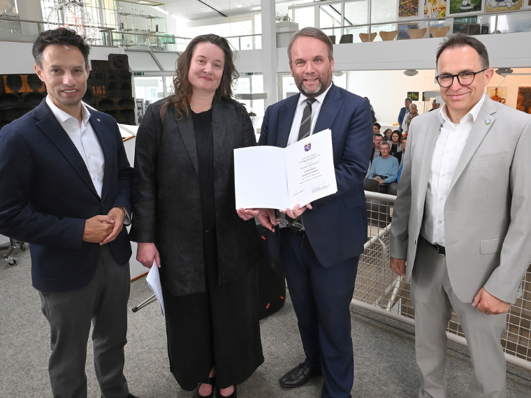 Vier Personen stehen in einem hellen Museumsfoyer bei einer offiziellen Übergabe. Ein Mann im blauen Anzug hält eine geöffnete Urkunde in die Kamera, flankiert von drei weiteren Personen in Businesskleidung. Im Hintergrund sind Sitzpublikum, Ausstellungsräume und Kunstwerke zu sehen.
