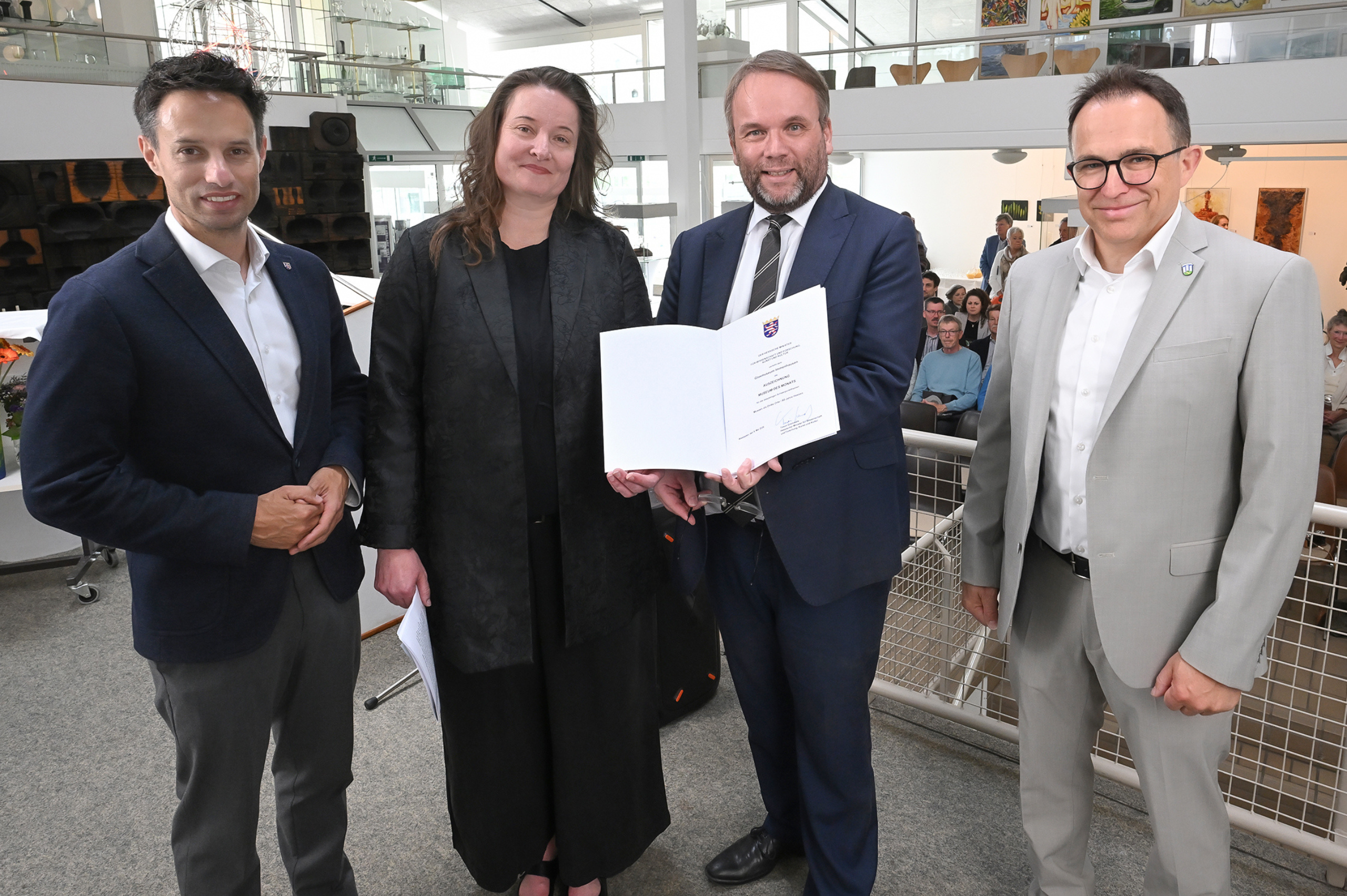 Vier Personen stehen in einem hellen Museumsfoyer bei einer offiziellen Übergabe. Ein Mann im blauen Anzug hält eine geöffnete Urkunde in die Kamera, flankiert von drei weiteren Personen in Businesskleidung. Im Hintergrund sind Sitzpublikum, Ausstellungsräume und Kunstwerke zu sehen.