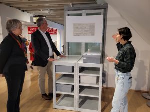 Drei Personen stehen vor einem Depotregal mit Kartons. Eine Frau erläutert anhand eines Auszugs aus einem früheren Inventarbuch die damalige Depotverwaltung.