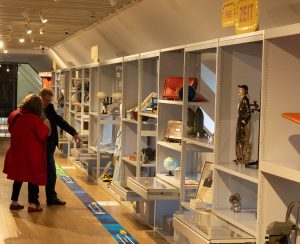 Museumsgäste stehen im Museum vor einer langen Wandvitrine, in der verschiedene Objekte gezeigt werden.
