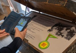 Hände richten ein Smartphone auf eine Holztruhe. Darin befindet sich eine Text-Bild-Tafel mit einem QR-Code.