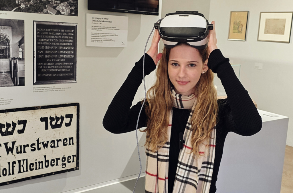 Person setzt in einer Ausstellung ein Virtual-Reality-Headset auf. Im Hintergrund befinden sich Informationstafeln und Exponate.