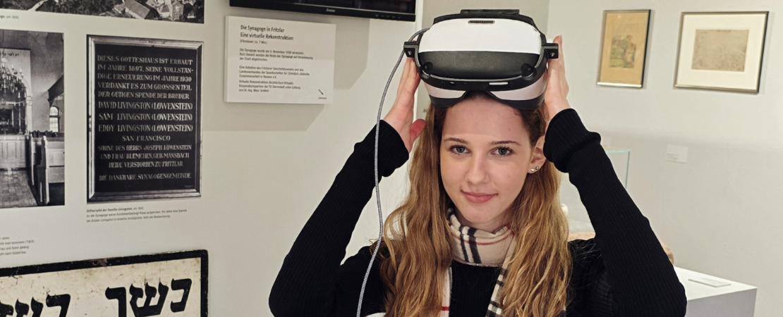 Person setzt in einer Ausstellung ein Virtual-Reality-Headset auf. Im Hintergrund befinden sich Informationstafeln und Exponate.