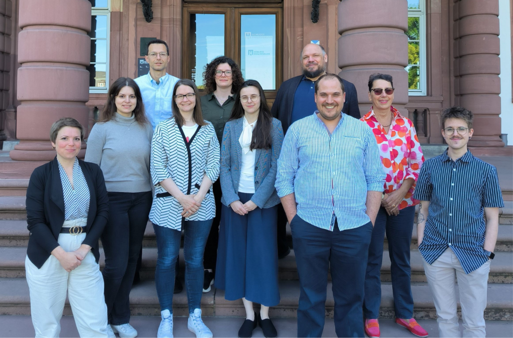 Gruppenfoto von zehn Personen vor einem historischen Gebäude mit Sandsteinfassade und breiter Treppe. Es handelt sich um das Historische Museum Schloss Phillipsruhedem in Hanau. Die Gruppe besteht aus sieben Absolvent*innen und die Veranstalter*innen des Kurses. Alle stehen auf den Stufen und blicken freundlich in die Kamera.
