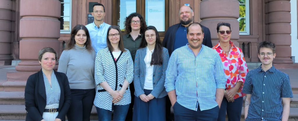 Gruppenfoto von zehn Personen vor einem historischen Gebäude mit Sandsteinfassade und breiter Treppe. Es handelt sich um das Historische Museum Schloss Phillipsruhe in Hanau. Die Gruppe besteht aus sieben Absolvent*innen und die Veranstalter*innen des Kurses. Alle stehen auf den Stufen und blicken freundlich in die Kamera.