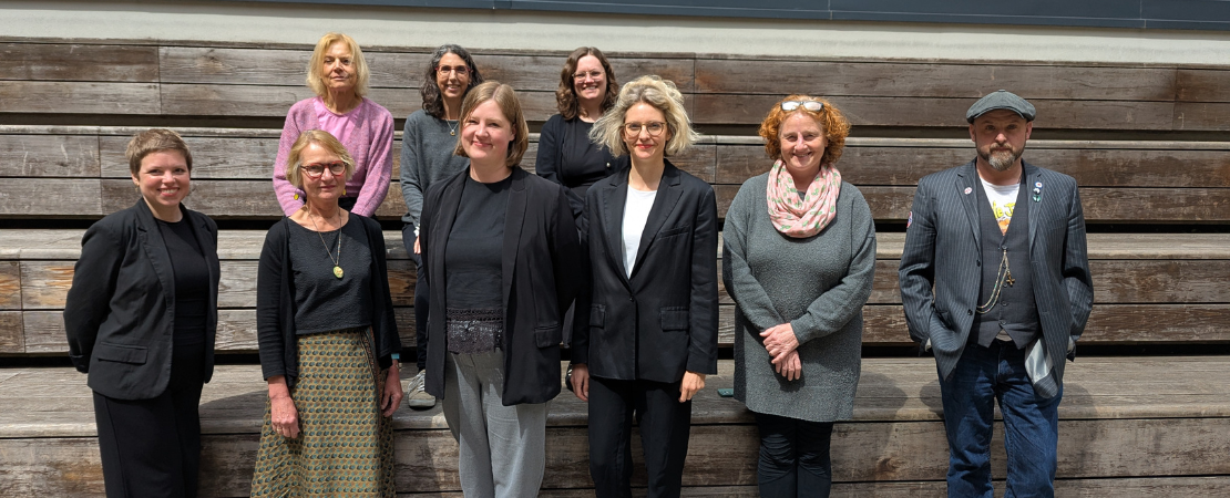 Gruppenfoto von neun Personen vor einer einer hölzernen Tribüne. Die Gruppe besteht aus Absolvent*innen und Verantwortlichen des Kurses. Alle blicken in die Kamera.