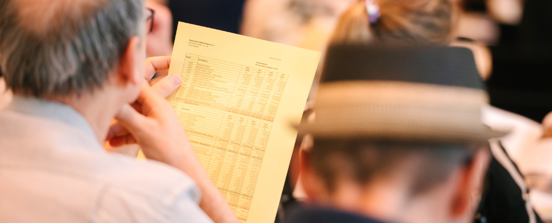 Mann in formaler Kleidung, der konzentriert ein Papier, vermutlich den Haushaltsplan, bei der Mitgliederversammlung liest.