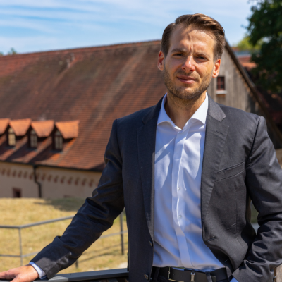 Porträt einer männlichen Person in dunkelblauem Anzug und weißem Hemd. Sie lehnt an einem Geländer und blickt in die Kamera. Im Hintergrund sind ein Gebäude mit rotem Ziegeldach und ein sonniger Himmel zu sehen.