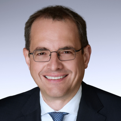 Portraet einer männlichen Person in Jackett mit weißem Hemd und blauer Krawatte. Er lächelt in die Kamera und trägt eine Brille.