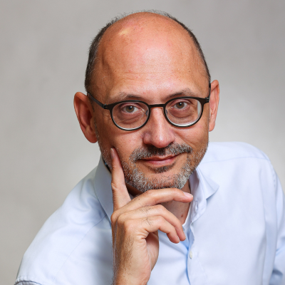 Portrait einer männlichen Person die in die Kamera schaut. Er trägt eine Brille und stützt sein Kinn auf der Hand ab.