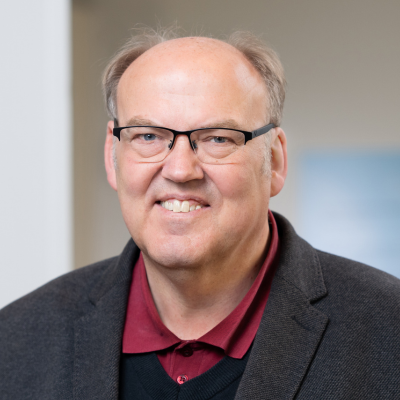 Portrait von einer männlichen Person, die in die Kamera schaut. Er trägt eine Brille und ein graues Jackett mit schwarzer Weste und rotem Hemd darunter.