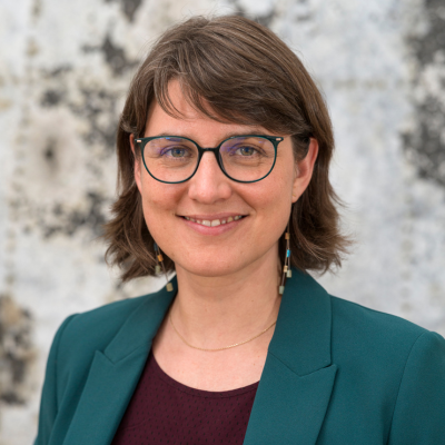 Porträt einer lächelnden Person mit kurzem braunem Haar und runder Brille. Sie trägt ein petrolfarbenen Blazer über einem roten Oberteil sowie schlichte, hängende Ohrringe. Der Hintergrund ist hell und unscharf.