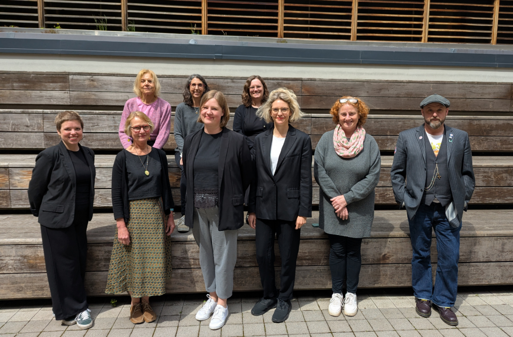 Gruppenfoto von neun Personen vor einer einer hölzernen Tribüne. Die Gruppe besteht aus Absolvent*innen und Verantwortlichen des Kurses. Alle blicken in die Kamera.