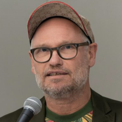 Person mit Brille, kurzem Bart und gemusterter Schildmütze, die in die Kamera blickt, vor ihr ein Mikrofon. Der Hintergrund ist grau.