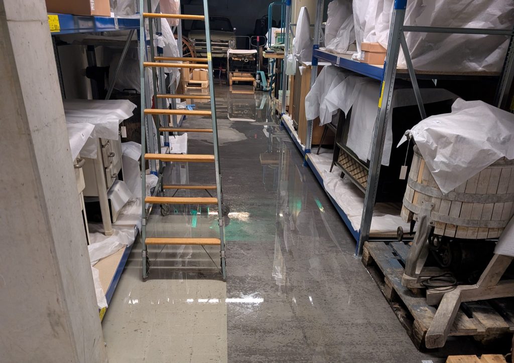 Depot mit Regalen auf der linken und rechten Seite, in denen verpackte Objekte stehen. In der Mitte steht eine Leiter. Auf dem Boden ist Wasser.