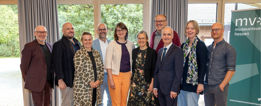 Gruppenfoto des Vorstands des Museumsverbands Hessen mit 10 Personen. Die Gruppe steht in einem hellen Raum vor großen Fenstern und einem Banner des Museumsverbands Hessen. Die Personen tragen unterschiedliche Kleidung und blicken in die Kamera.