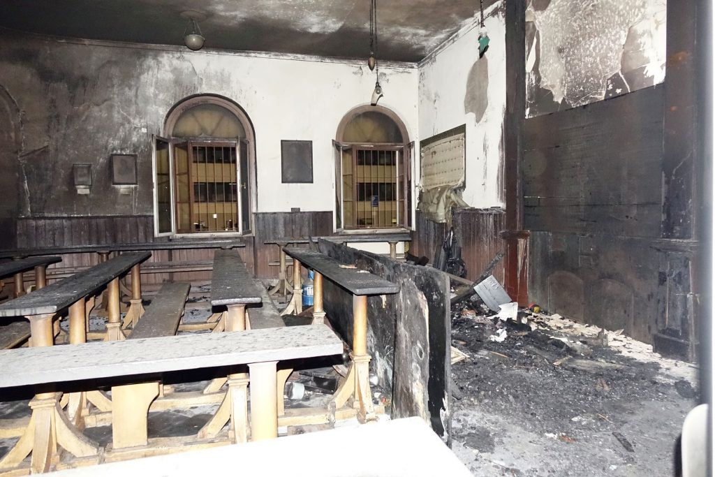 Innenraum mit Brandschäden: verkohlte Holzbänke und Tische, verrußte Wände und Brandschutt auf dem Boden.