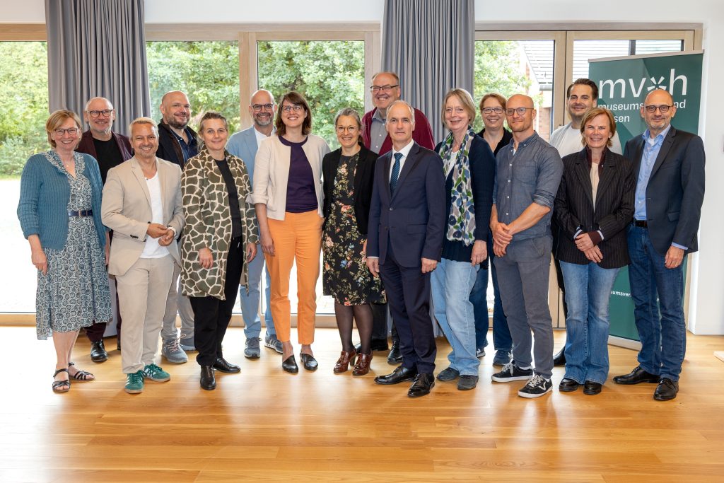 Gruppenfoto des Vorstands des Museumsverbands Hessen mit 16 Personen. Die Gruppe steht in einem hellen Raum vor großen Fenstern und einem Banner des Museumsverbands Hessen. Die Personen tragen unterschiedliche Kleidung und blicken in die Kamera.