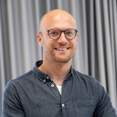 Lächelnde Person mit Brille. Sie trägt ein dunkelgraues Hemd über einem weißen Oberteil und blickt direkt in die Kamera. Der Hintergrund ist unscharf.