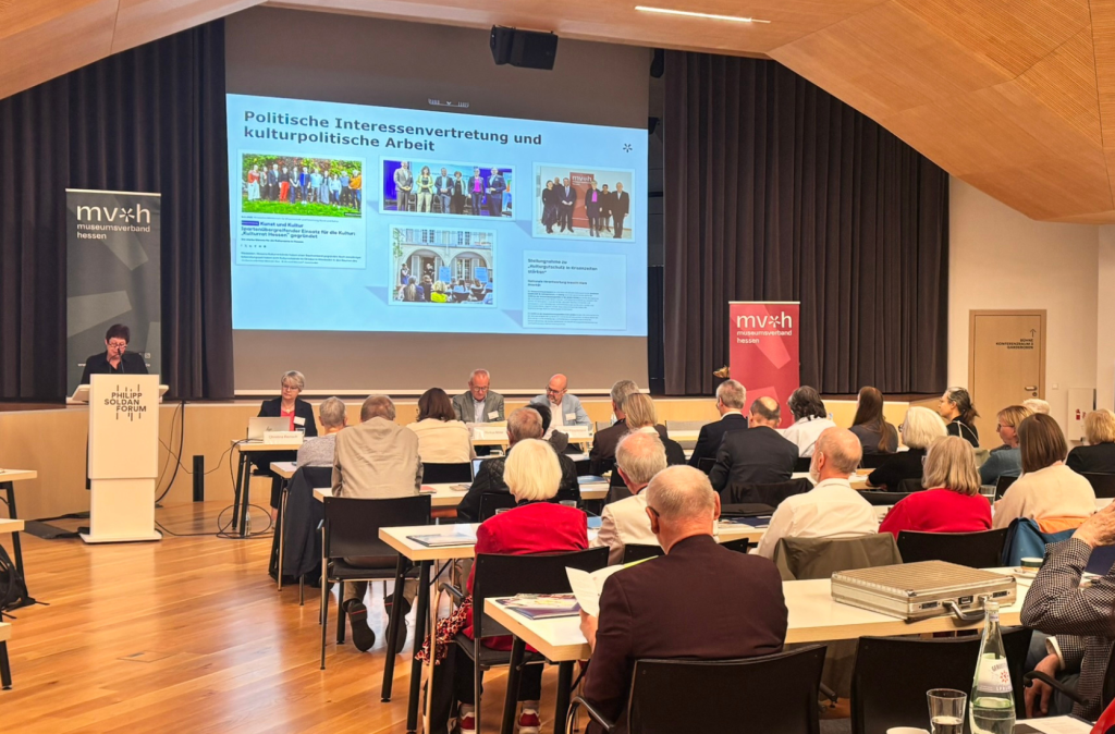 Teilnehmende sitzen an Tischen und verfolgen einen Vortrag. Auf der Leinwand wird eine Präsentation zum Thema Politische Interessenvertretung und kulturpolitische Arbeit angezeigt.