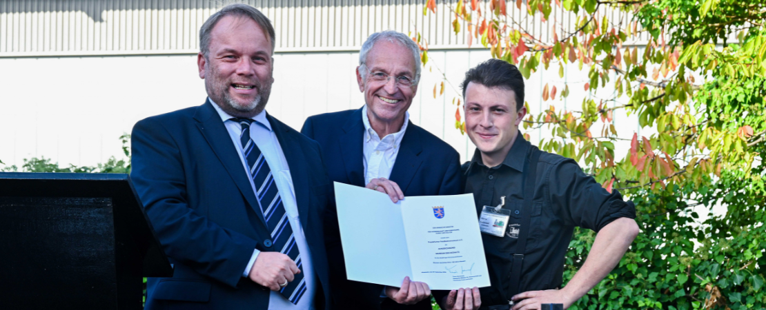 Drei Männer stehen nebeneinander. Alle drei lächeln. Der mittlere Mann hält eine geöffnete Urkunde in die Kamera.