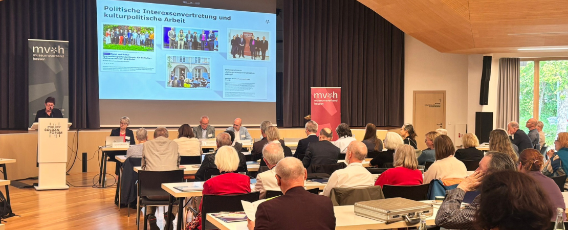 Teilnehmende sitzen an Tischen und verfolgen einen Vortrag. Auf der Leinwand wird eine Präsentation zum Thema Politische Interessenvertretung und kulturpolitische Arbeit angezeigt.