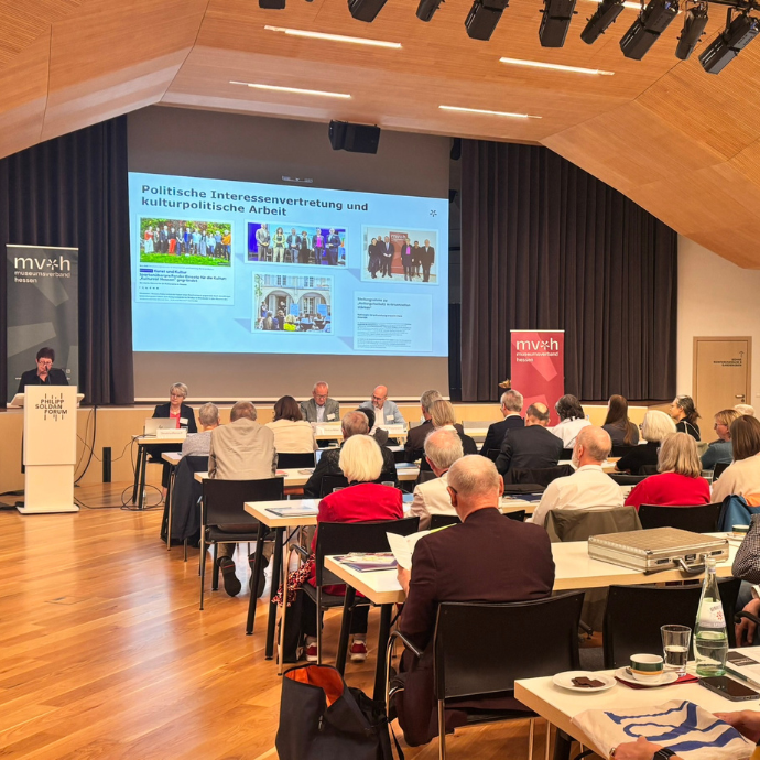 Teilnehmende sitzen an Tischen und verfolgen einen Vortrag. Auf der Leinwand wird eine Präsentation zum Thema Politische Interessenvertretung und kulturpolitische Arbeit angezeigt.