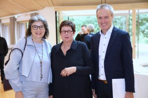 Drei Personen stehen lächelnd nebeneinander in einem hellen Innenraum. Die Frau links trägt eine helle Jacke und eine bunte lange Halskette, die Frau in der Mitte eine schwarze Bluse und eine runde Brille. Rechts steht ein Mann im dunklen Anzug mit einem Dokument unter dem Arm.