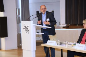 Eine Person mit Brille, dunklem Jackett und hellblauem Hemd steht am Rednerpult und spricht ins Publikum.