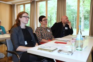 Drei Teilnehmende sitzen an einem Tisch und verfolgen aufmerksam einen Vortrag. Vor ihnen liegen Unterlagen, Notizbücher und Stifte. Die Veranstaltung findet in einem hellen Raum mit großen Fenstern und Blick ins Grüne statt.
