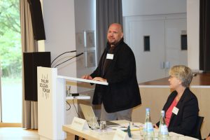 Eine Person mit einer schwarzen Jacke steht am Rednerpult und spricht ins Publikum. Eine weitere Person sitz daneben an einem Tisch. Auf dem Tisch stehen ein offener Laptop, Wasserflaschen und Dokumente.