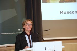 Eine Person mit Brille und kurzen Haaren steht am Rednerpult und spricht ins Publikum. Im Hintergrund ist ein Aufsteller zu sehen. Eine Leinwand ist neben dem Aufsteller zu sehen.