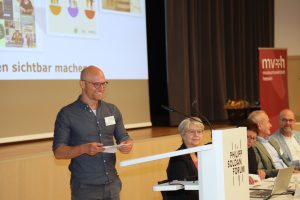 Eine Person mit Brille und Jeanshemd geht zu einem Rednerpult. Hinter dem Rednerpult sitzen weitere Personen an einem Tisch.