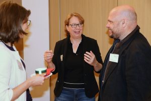 Drei Personen in einem Gespräch. Die Person in der Mitte spricht animiert mit den Händen zur Person rechts. Eine weitere Person steht mit Tasse daneben und verfolgt die Unterhaltung.