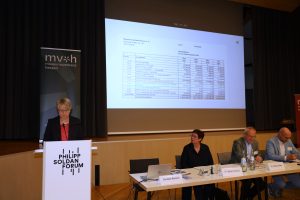 Eine Person steht am Rednerpult und spricht ins Publikum. Weitere Personen sitzen neben dem Pult an einem Tisch. Im Hintergrund ist ein Aufsteller vom Museumsverband Hessen und eine Leinwand zu sehen.