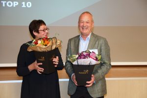Zwei Personen halten Blumensträuße in den Händen. Die linke Person schaut die andere Person an, während diese in die Kamera lacht.
