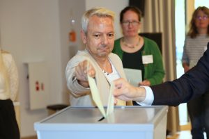 Eine Person in heller Kleidung gibt seinen Stimmzettel in eine Wahlurne. Daneben wirft eine weitere Person ihren Stimmzettel in die Wahlurne. Im Hintergrund sind weitere Personen unscharf zu sehen.