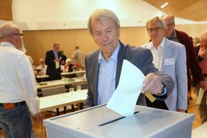 Eine Person in grauem Jackett gibt seinen Stimmzettel in eine Wahlurne. Im Hintergrund sind weitere Personen zu sehen.